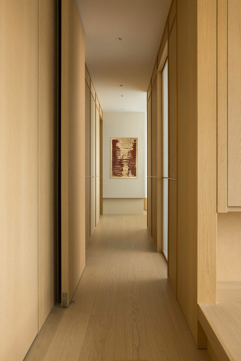 A warmly lit minimalist corridor with wooden panels and floors, leading to a framed abstract painting at the end.