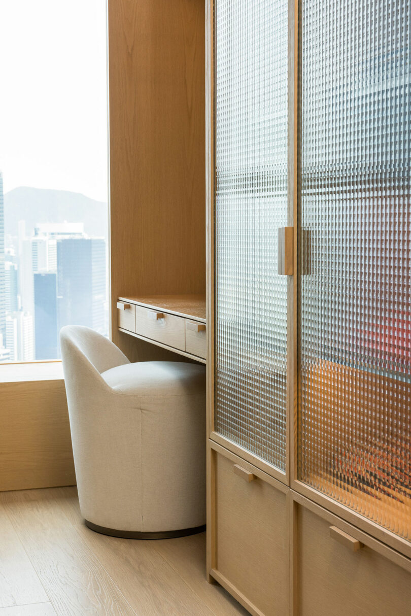 A modern office corner with a cream-colored armchair beside a wooden cabinet, overlooking a cityscape through a patterned glass window.