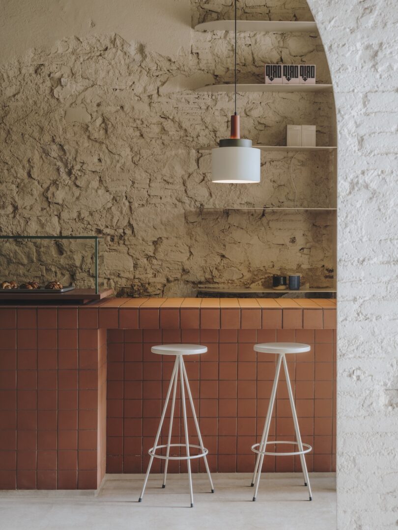 A rustic cafe interior with a textured stone wall, a counter made of square red tiles, two white metal stools, a hanging light fixture, and a shelf with a few items