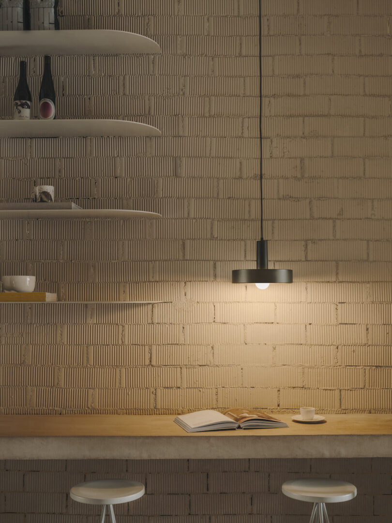 A pendant lamp hangs over a counter with an open book and a cup. Shelves with bottles and dishes are mounted on a textured brick wall