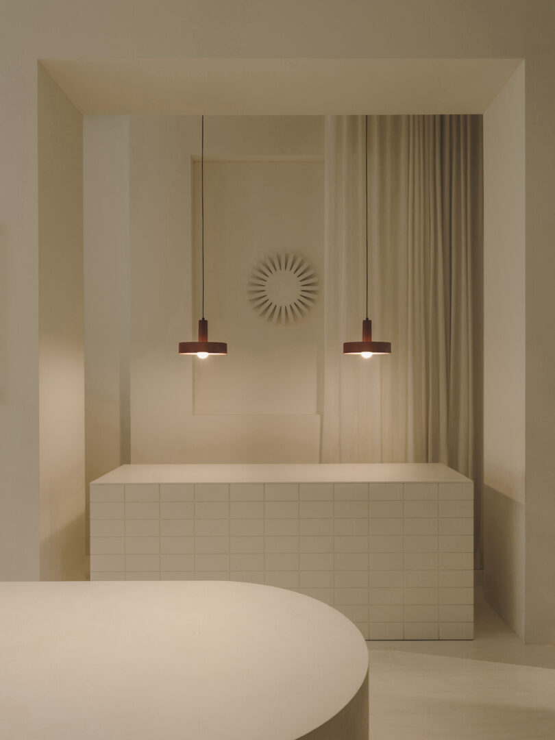 Minimalist interior with two pendant lights above a white tiled counter. A circular wall decor and beige curtain are in the background. A rounded countertop is visible in the foreground