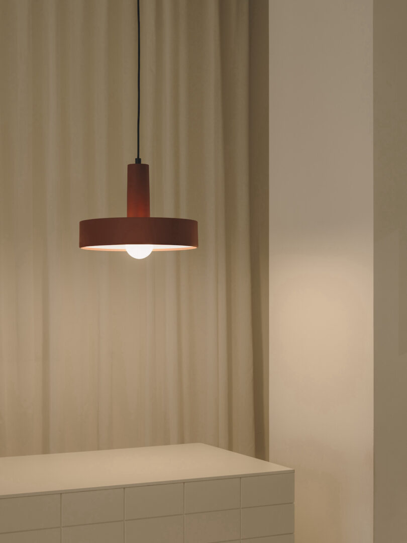 A hanging light fixture with a red shade is turned on above a white tiled counter, set against a beige curtain backdrop