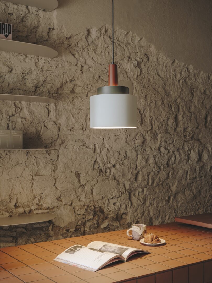 A cozy corner with a book and cup of coffee on a tiled surface, illuminated by a modern pendant light, against a textured stone wall with shelves