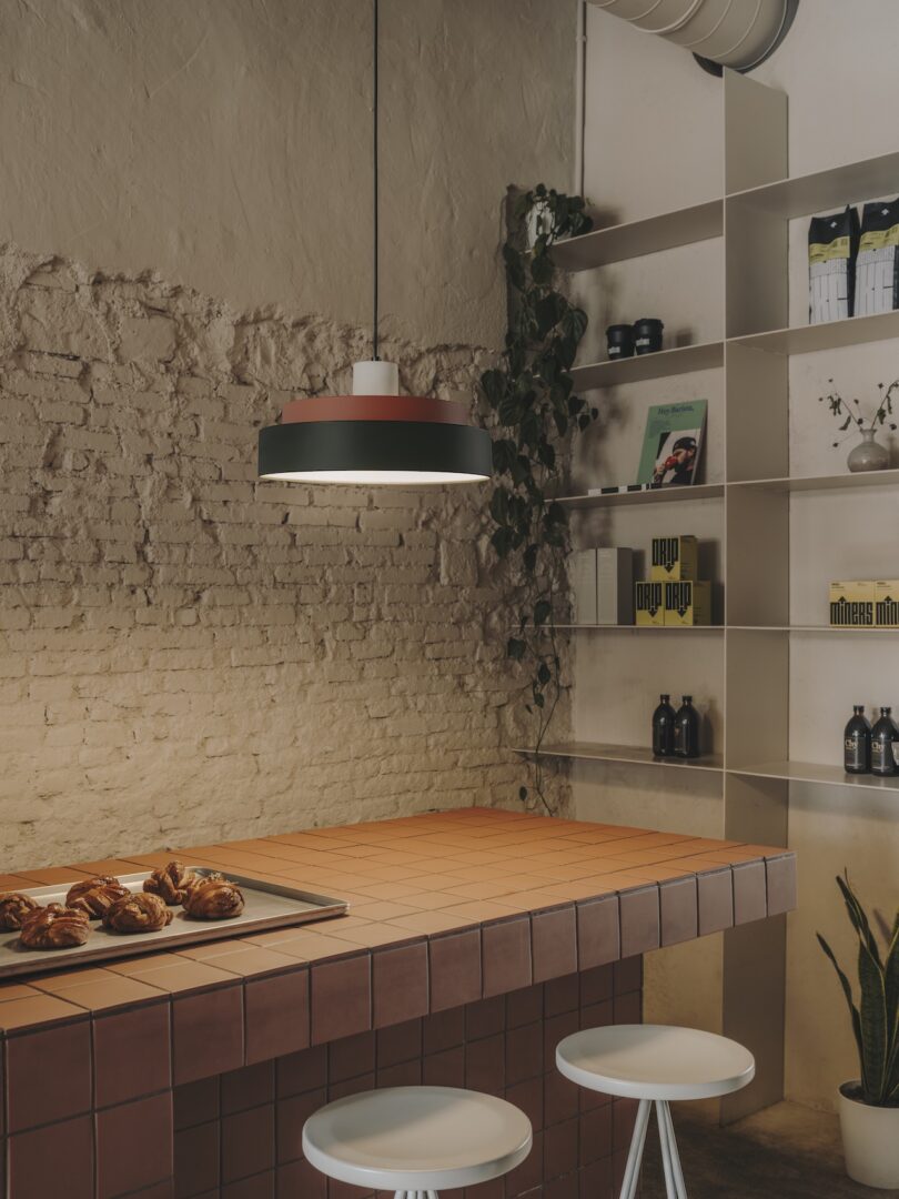 A kitchen corner with a brick wall, a countertop with pastries, three white stools, a hanging light fixture, and shelves with various items including books, plants, and bottles