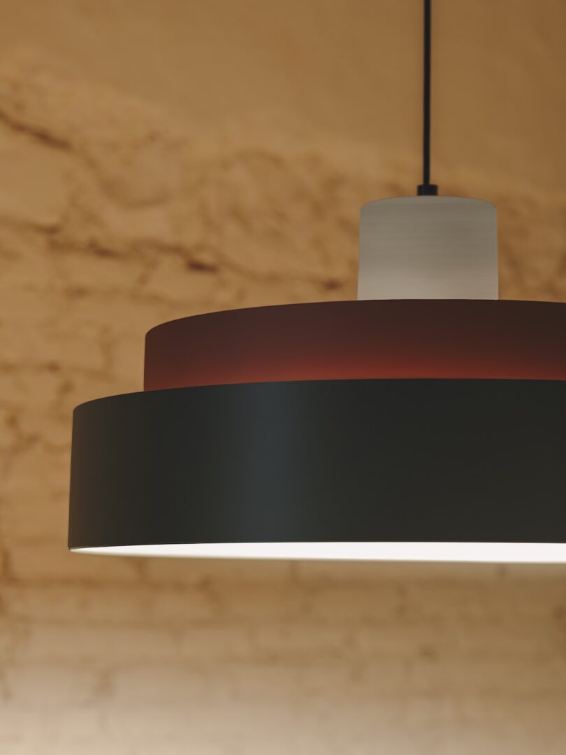 Close-up of a modern ceiling light fixture with a black and red shade, hanging against a beige brick wall background