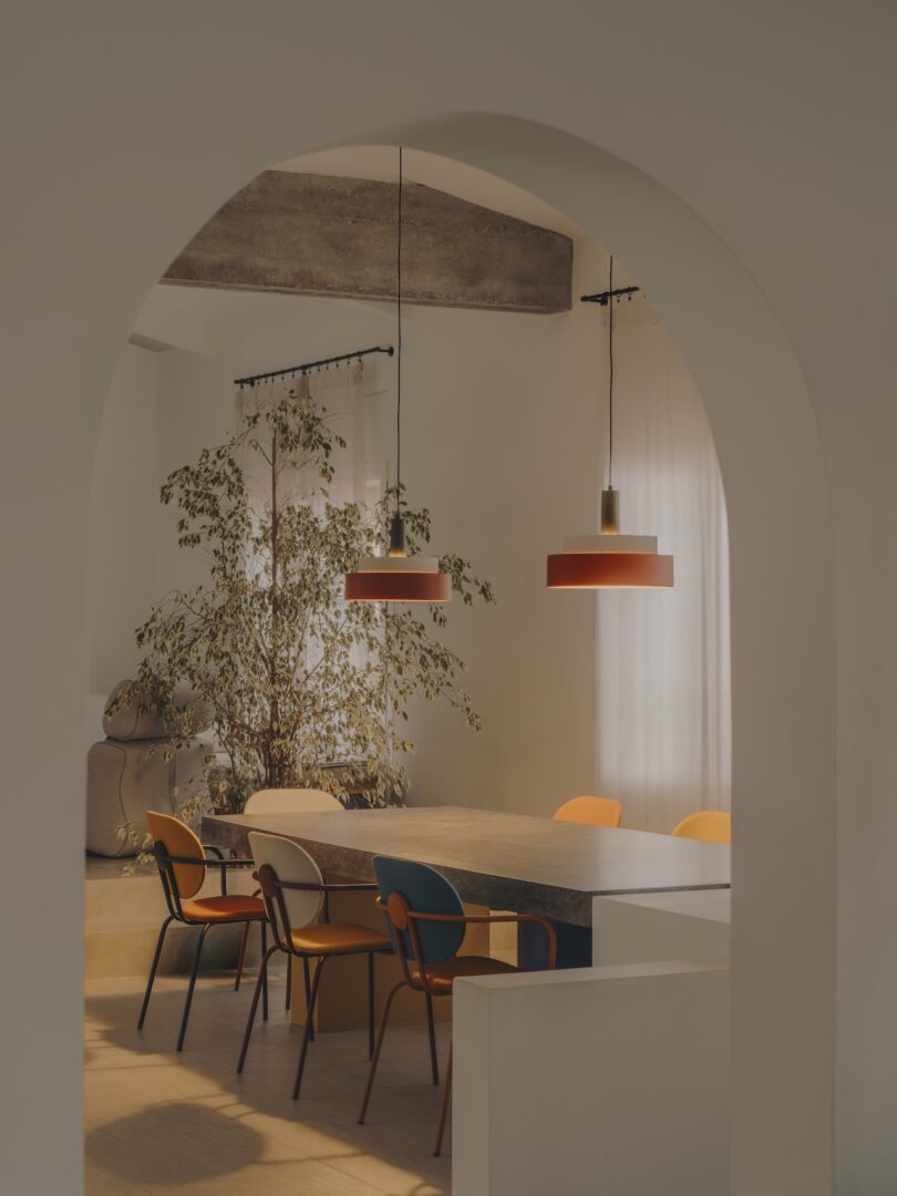 A dining area viewed through an archway, featuring a table with multicolored chairs, two pendant lights above, and a large indoor plant in the background