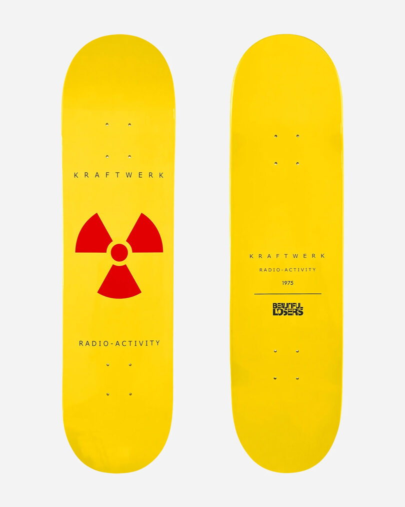 Two yellow skateboards from the Kraftwerk Collection. One features a red radioactive symbol with the text "Kraftwerk Radio-Activity," while the other showcases "Kraftwerk Radio-Activity 1975" and "Bruisers Losers." 