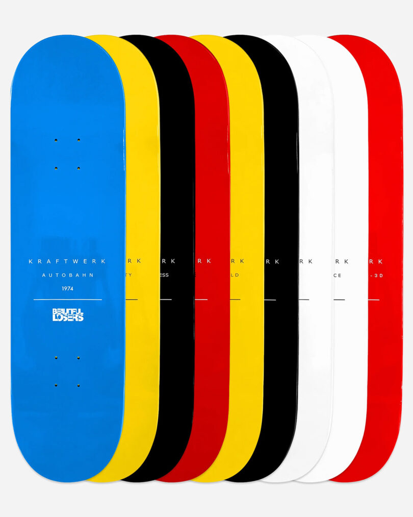 A collection of nine colorful skateboard decks from the Kraftwerk Collection displayed in a row, featuring blue, yellow, red, black, and white colors. Each deck is marked with text and graphics from the Kraftwerk album catalog.