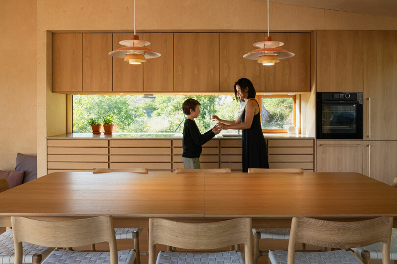 A woman and a child stand in a modern kitchen with wooden cabinets and a large dining table, lit by two pendant lights.