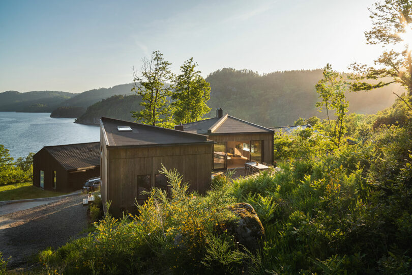 Modern wooden house situated on a lush hillside overlooking a serene lake and surrounded by trees and greenery with mountains in the background.