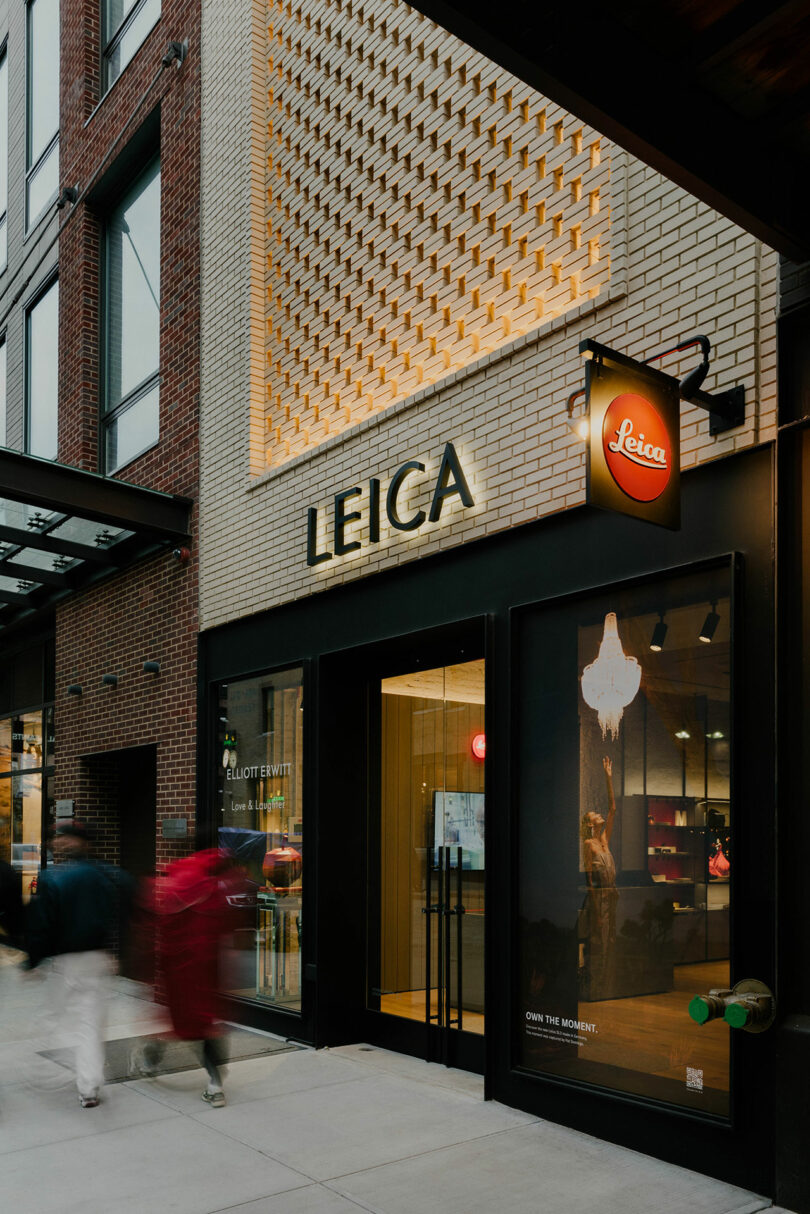 Exterior of a leica camera store at twilight with blurred figures passing by, showcasing the brand's illuminated signage and display windows.