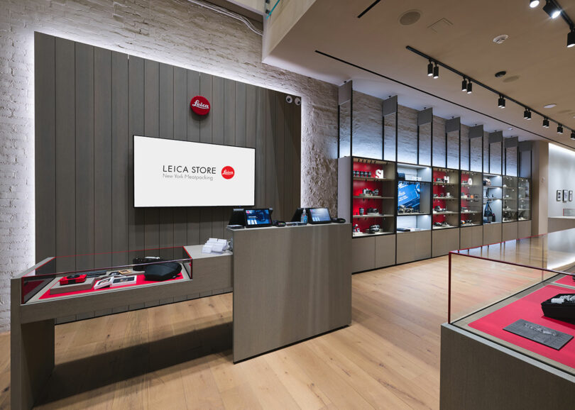 Interior of a leica camera store showing display cases with cameras and lenses, a digital screen, and a branded wall behind the counter.