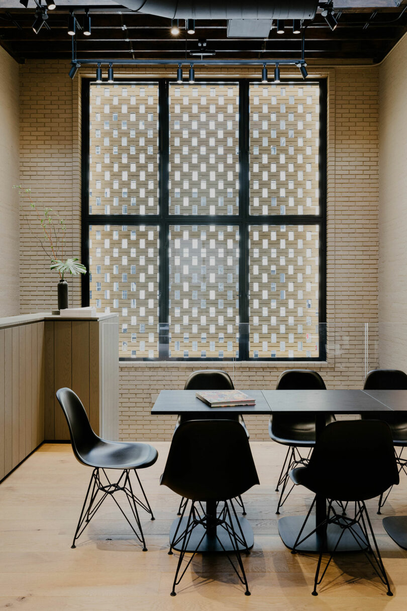 Modern office meeting room featuring a long table with black chairs, large windows with a patterned glass design, and a textured beige brick wall.