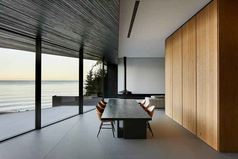 Modern dining room in a Liminal House with wooden walls and a large table, expansive glass windows offering a view of the ocean, and a detailed textured ceiling.