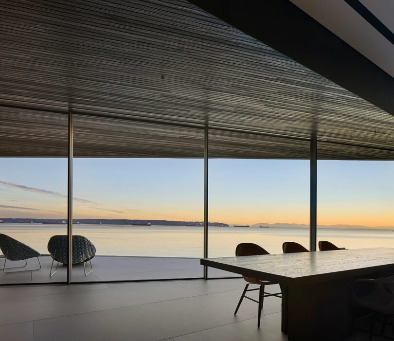 Liminal House interior with a large wooden table and two chairs overlooking a tranquil sea view through floor-to-ceiling windows at sunset.