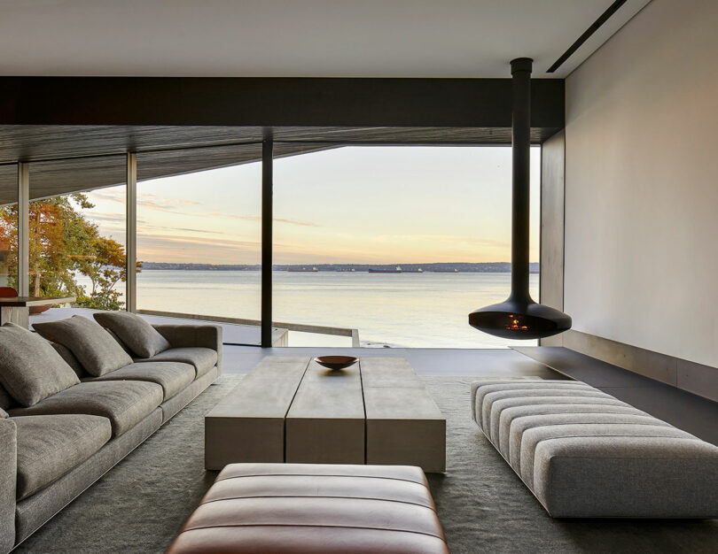 Liminal House living room with large windows overlooking a lake, featuring a suspended fireplace, grey sectional sofa, and ottomans.