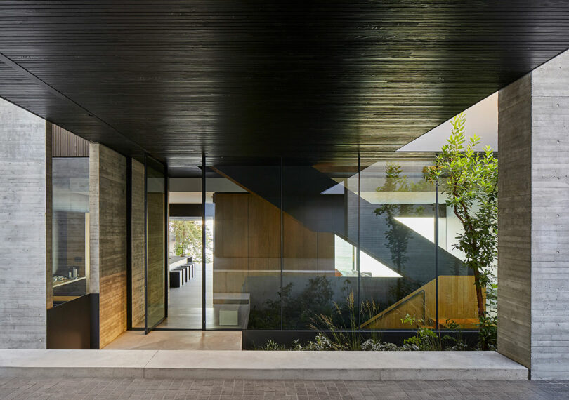covered patio exterior of a modern concrete house looking through wall of glass into large home with black staircase.