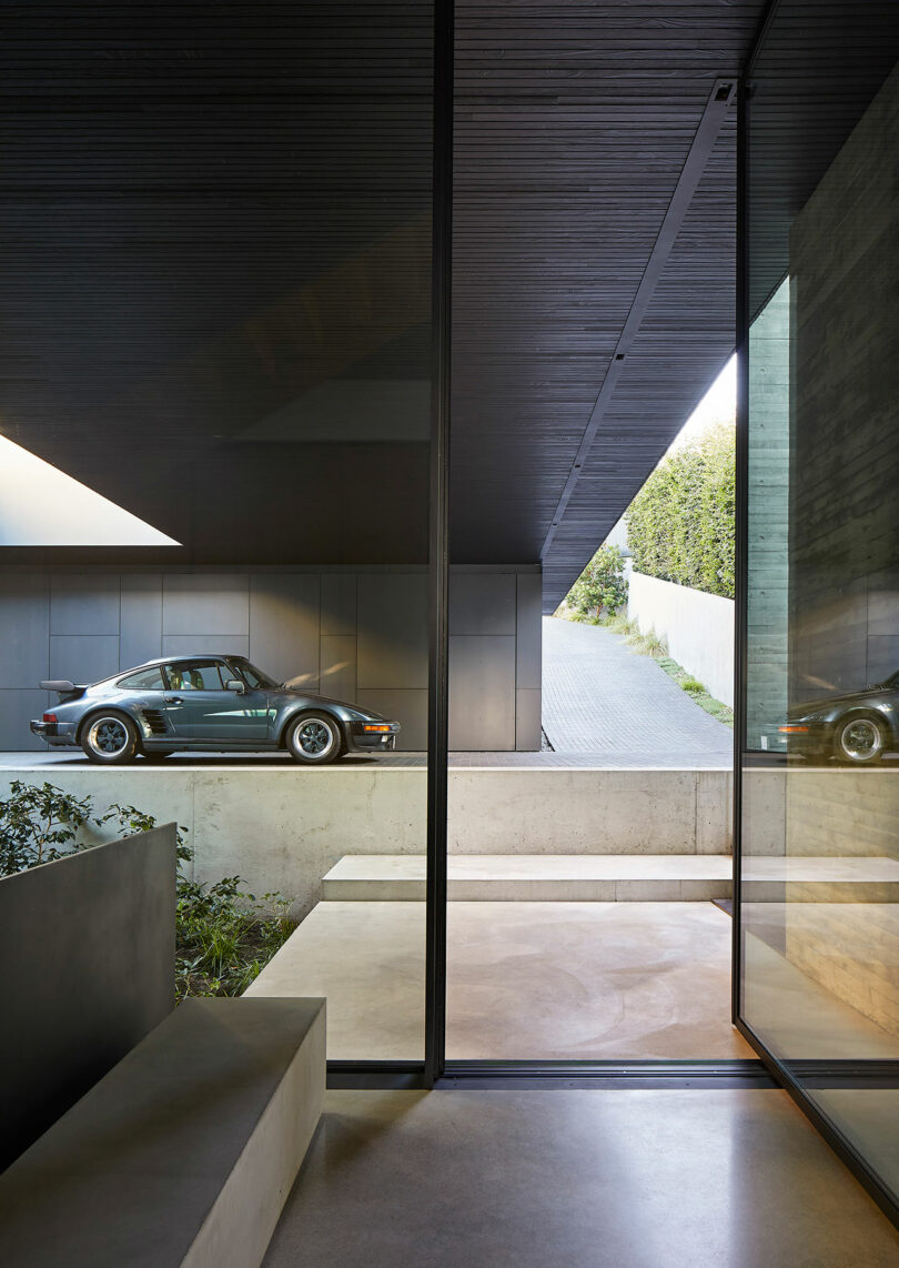 A vintage car reflected in the large glass window of a liminal house with minimalist architectural features and concrete flooring.