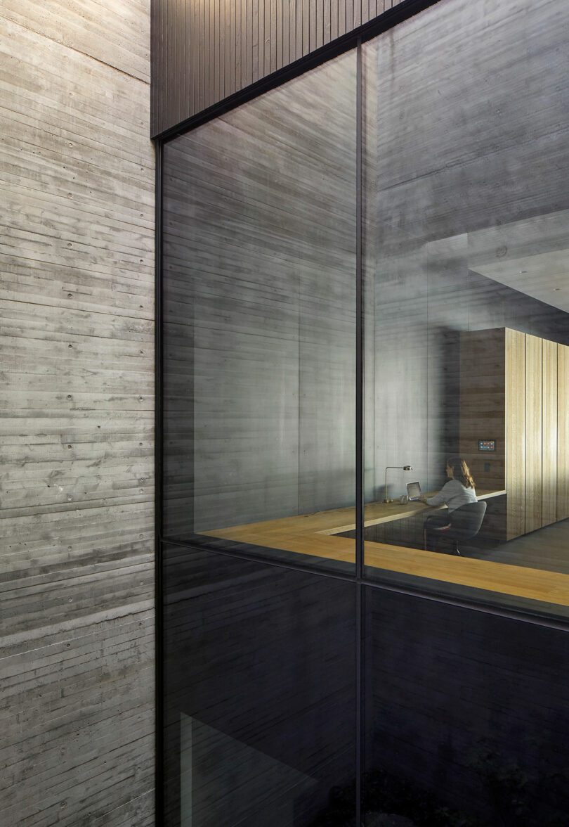 Interior courtyard of modern concrete house looking through glass windows at woman working at desk.