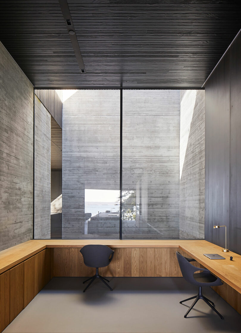 Modern office interior with wooden desks, black chairs, and large glass windows overlooking a liminal house.