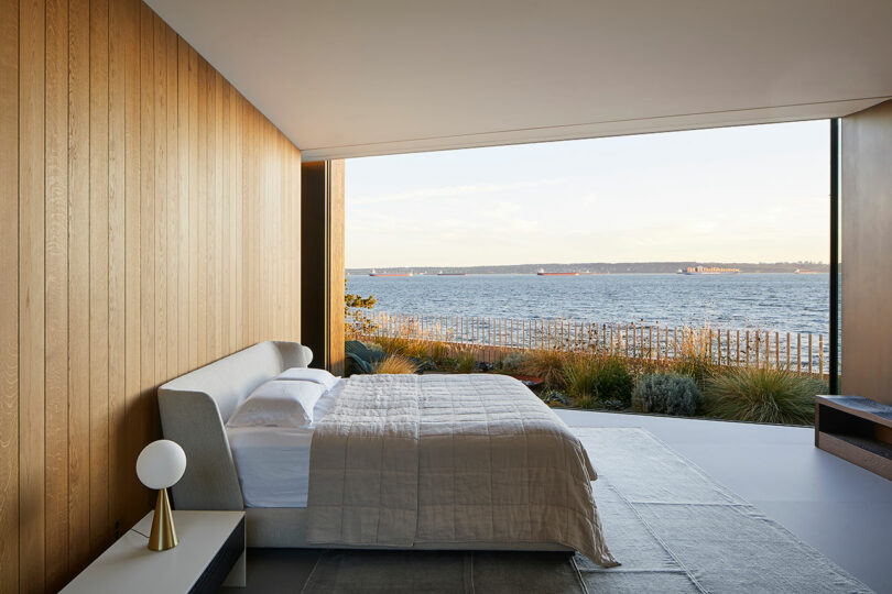 Modern bedroom in a Liminal House with large window overlooking the ocean, featuring a minimalist bed, wooden walls, and a bedside lamp.