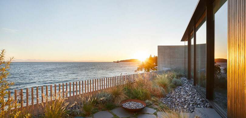 Sunset view from a modern coastal Liminal House with large windows, wooden fence, and landscaped garden featuring stones and shrubs.