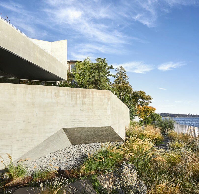 A modern concrete house with geometric design elements overlooking a coastal landscape with lush greenery and a clear sky.