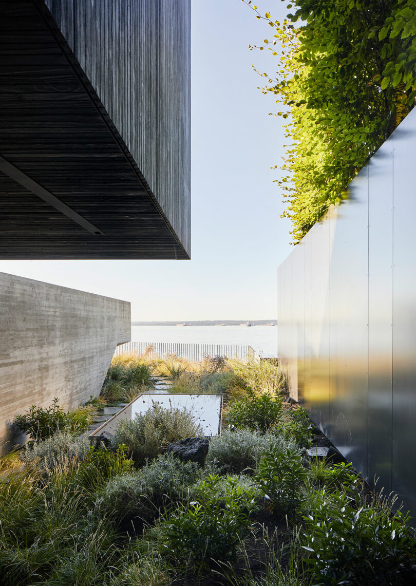 A modern architectural garden featuring lush greenery, water features, and concrete elements, with a view of the House beside a distant water body under a clear sky.