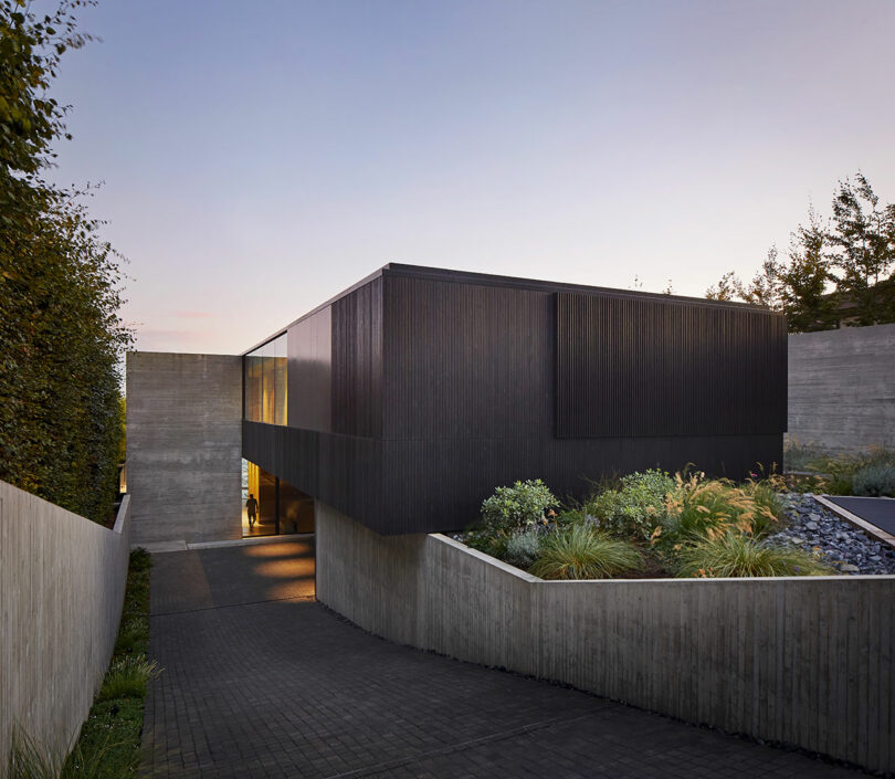 Angled front view of boxy modern house made of concrete and black wood with front garden.
