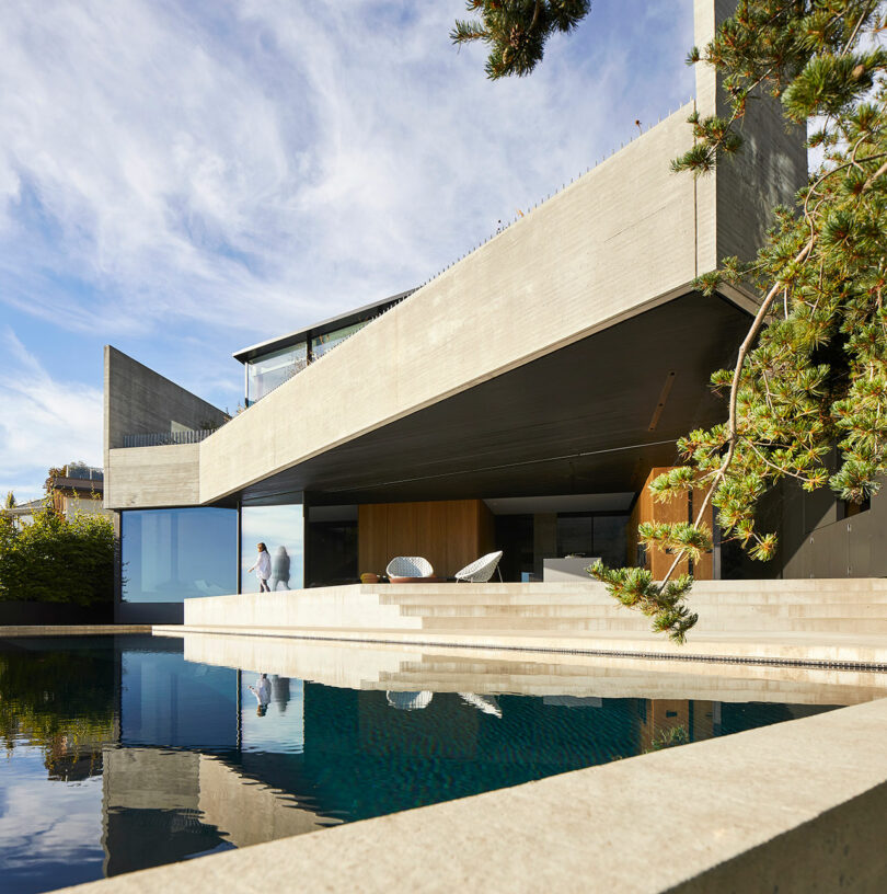 Liminal house featuring large windows and wood paneling, with a reflective pool in the foreground and a person walking inside.