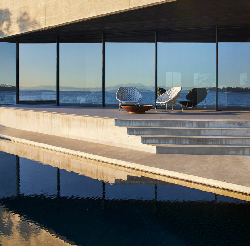 Modern chairs beside an infinity pool with clear glass windows at the Liminal House overlooking a serene lake at sunset.
