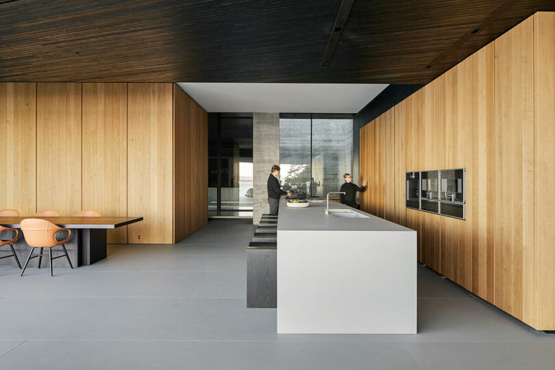 Liminal house interior with wooden walls and a minimalistic design, featuring a central island and two people, one at the counter and the other walking.