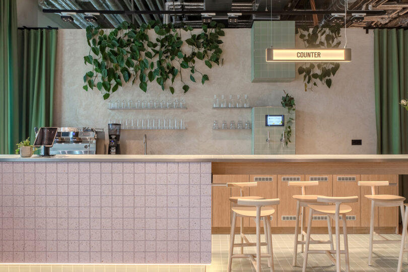 Modern cafe counter with tiled front and wooden bar stools; overhead sign reads "COUNTER." A coffee machine, hanging pots, and glasses are visible, with plants decorating the walls and ceiling. The ambiance feels like a stylish hotel lobby, reminiscent of the vibe at MM:NT Berlin Lab.