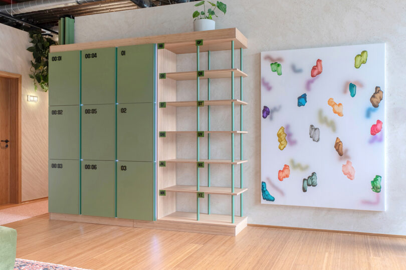 Communal locker units within the MM:NT Berlin Lab allowing guests for food delivery and other services. A large modern and colorful painting of floating nebulous shapes is wall mounted to the right of the locker shelving system.