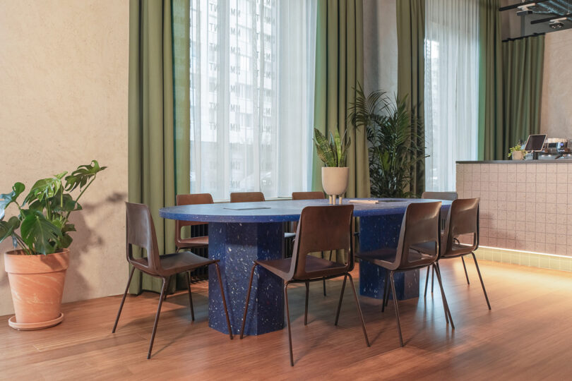 A modern communal dining area at the MM:NT Berlin Lab's Counter features a large blue oval table with eight brown chairs. The space boasts green curtains, potted plants, and ample natural light through tall windows, creating a refreshing ambiance akin to a stylish hotel lounge.