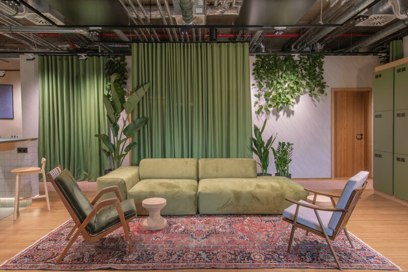 A modern room at the MM:NT Berlin Lab with green curtains, a matching green sofa, two chairs, plants, and a colorful rug on a wooden floor. A wooden door and cabinetry are in the background, creating an inviting hotel-like ambiance.