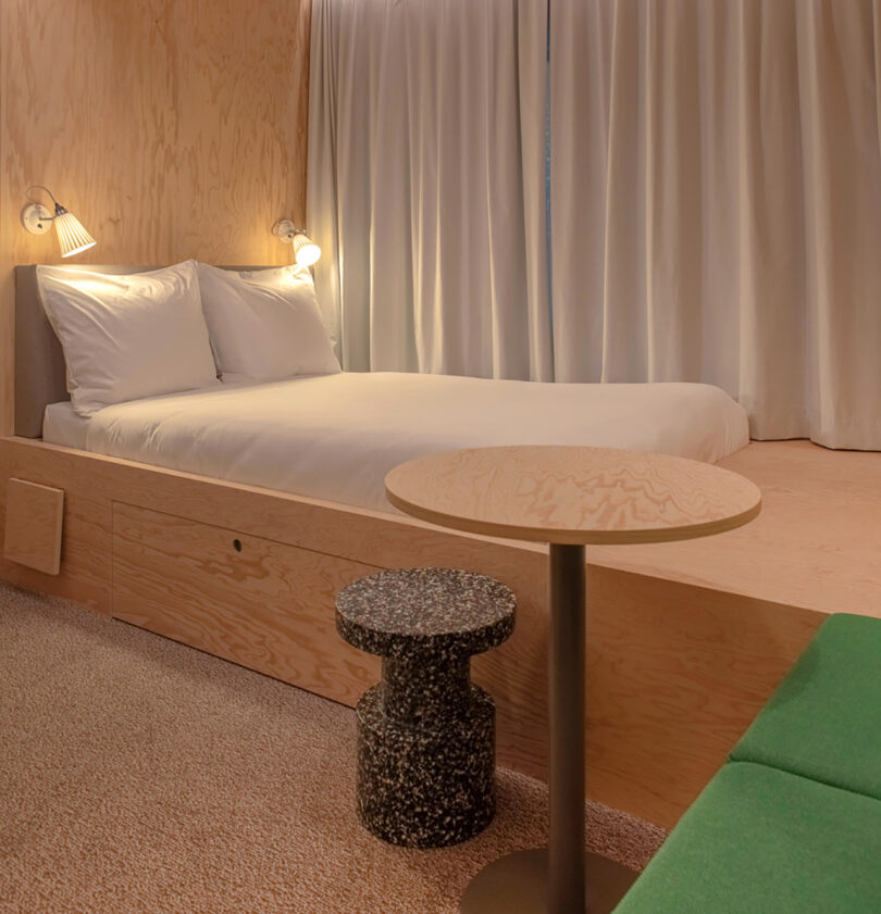 A minimalist bedroom in the MM:NT Berlin Lab hotel features a double bed on a wooden platform, two wall-mounted reading lights, a small round table with a textured stool, and green upholstered seating by the side.
