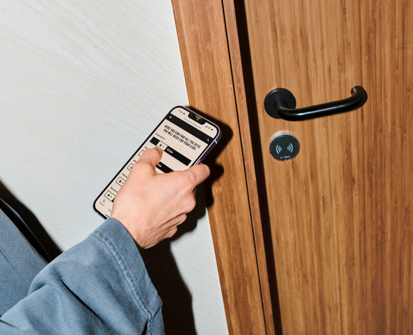 A person in a blue sleeve holds a smartphone near a door with an electronic lock, displaying a digital key or access screen at the beta testing MM:NT Berlin Lab hotel.