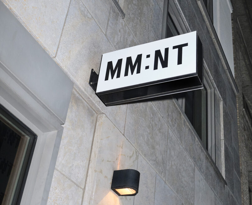 A wall-mounted sign on a building exterior reads "MM:NT" with a black and white color scheme, giving the appearance of a chic hotel entrance.