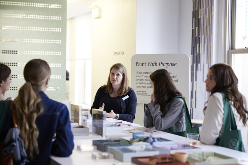 A woman presenting at a booth labeled "paint with purpose" at NeoCon to three listeners in a brightly lit room.