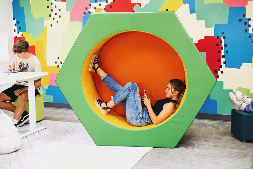 A woman lounges in a circular green and orange chair in a colorful room while others study at tables nearby.