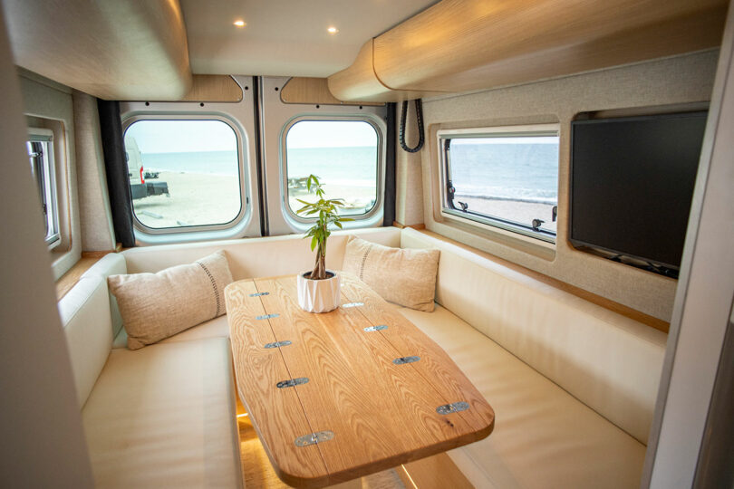 The image shows a cozy RV interior with a wooden table, surrounding white cushioned seating, a mounted TV, and a window view of a beach. A potted plant sits in the center of the table.