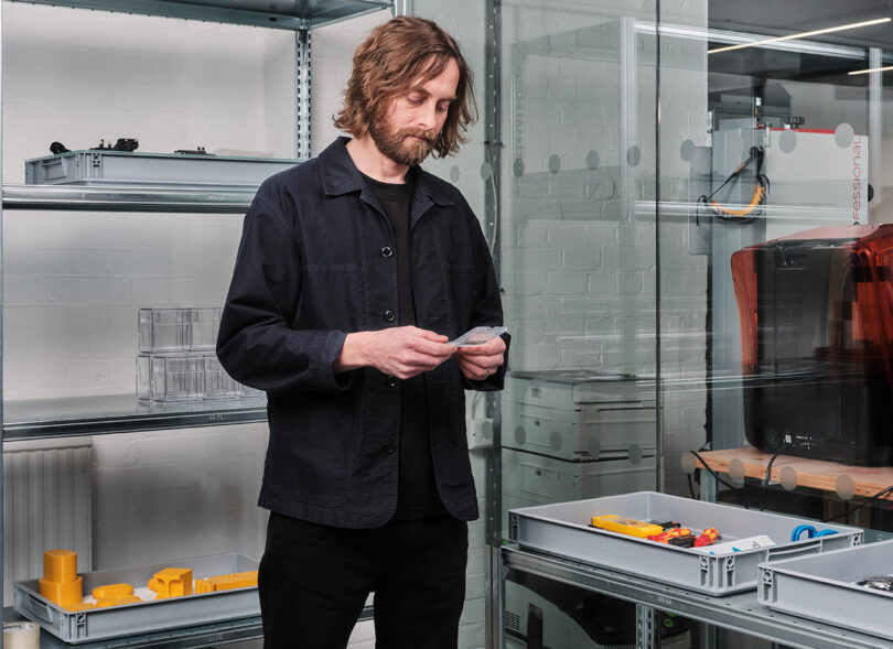 Adam Bates, Design Director at Nothing, with shoulder-length hair wearing a dark jacket examines a Nothing Phone (2a) Special Edition, in a room with shelves containing tools and components.