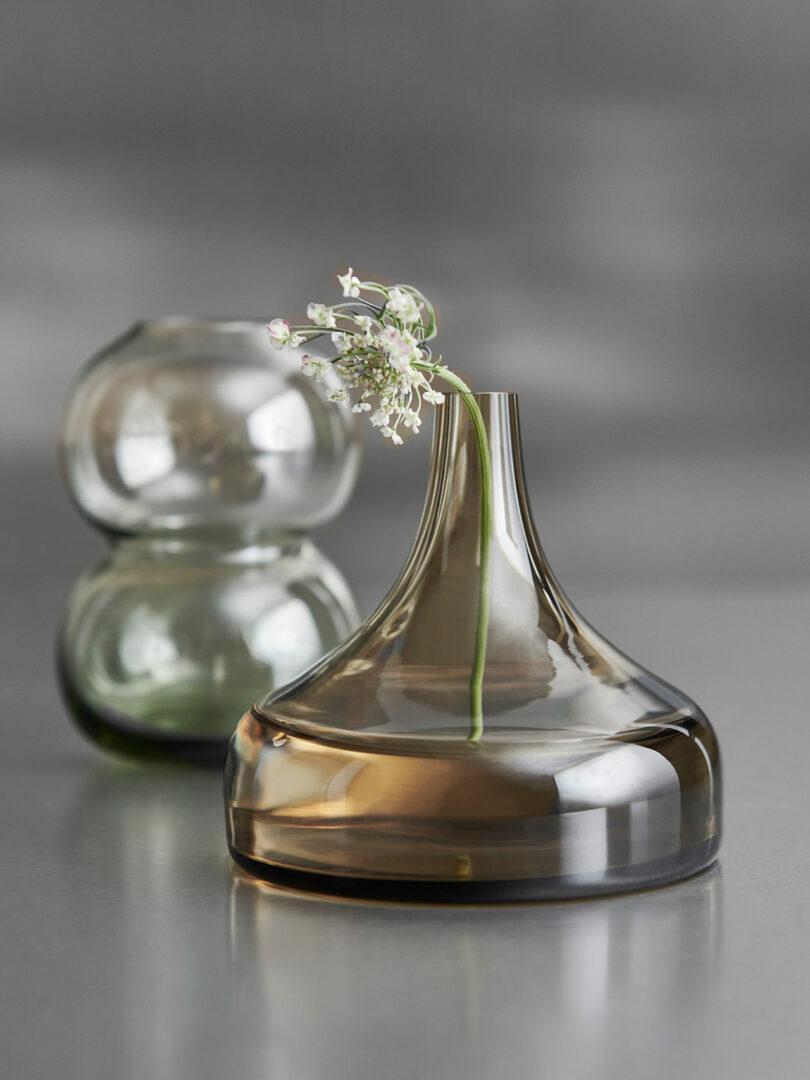 A translucent, brown vase holding a single white flower stands in the foreground, with a round, transparent object in the background on a smooth, gray surface.