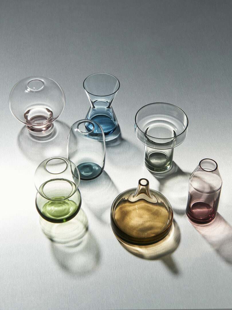 A collection of seven glass objects in various shapes and colors, including green, brown, pink, blue, and clear, arranged on a reflective surface.
