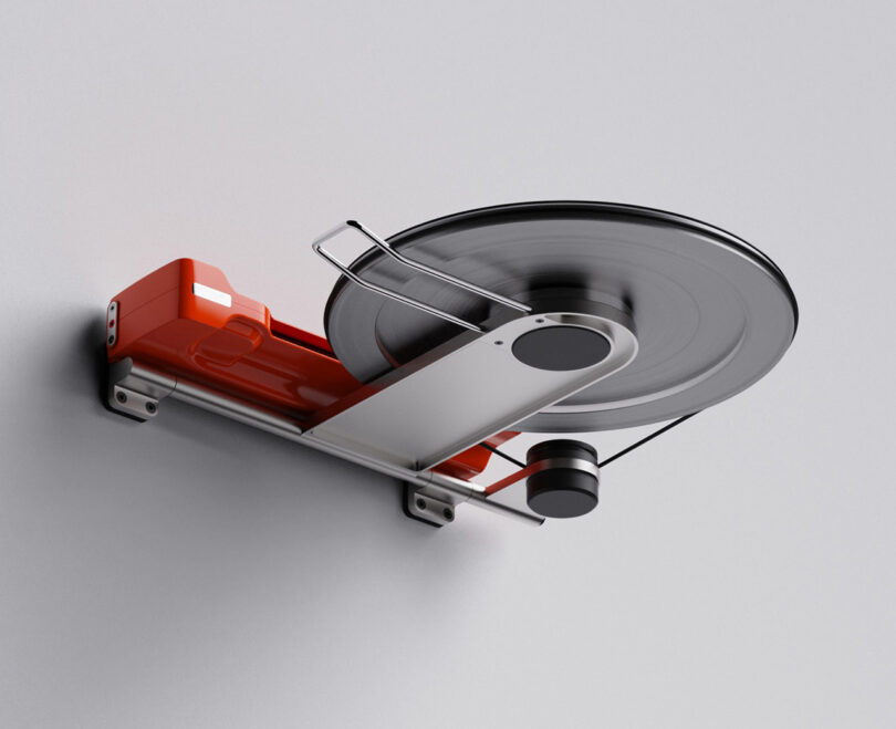 A modern, minimalist red and silver wall-mounted reel-to-reel record player viewed from underneath revealing a hinged platter arm