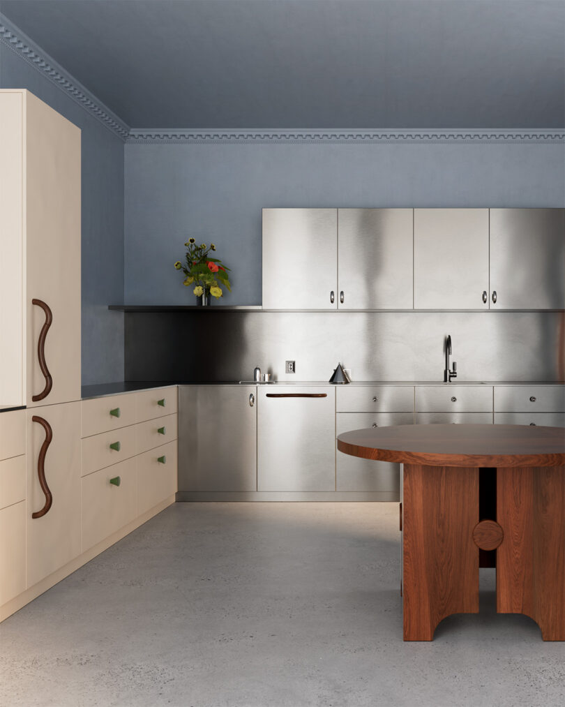 Modern kitchen with blue walls, stainless steel cabinets and appliances, beige cupboards with curved handles, a wooden table, and a vase with flowers on the counter.