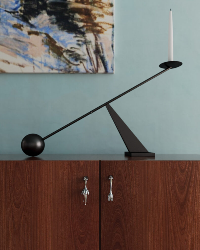 A minimalist candle holder with a black base and spherical counterweight sits on a wooden cabinet. A painting hangs on the light blue wall behind it.