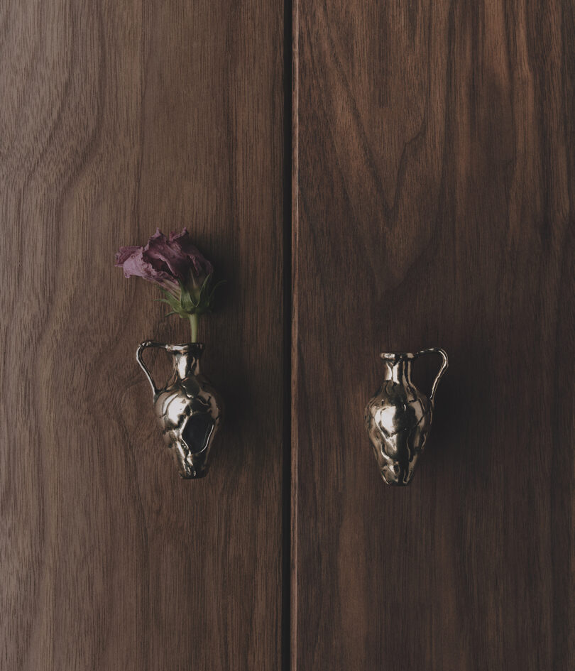 Wooden surface with two bronze-colored pulls shaped like pitchers; one holds a dried purple flower.