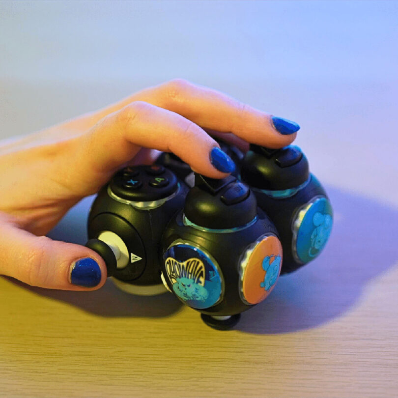 A hand with blue nail polish holding a black spherical fidget toy with buttons and colorful stickers, reminiscent of the design elements on a Proteus gaming controller.